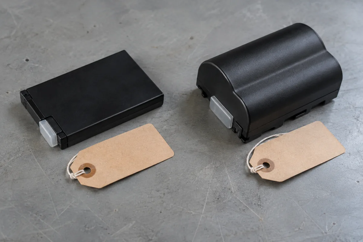 Flat rectangular battery pack and thicker curved battery pack beside blank tags on a gray bench.